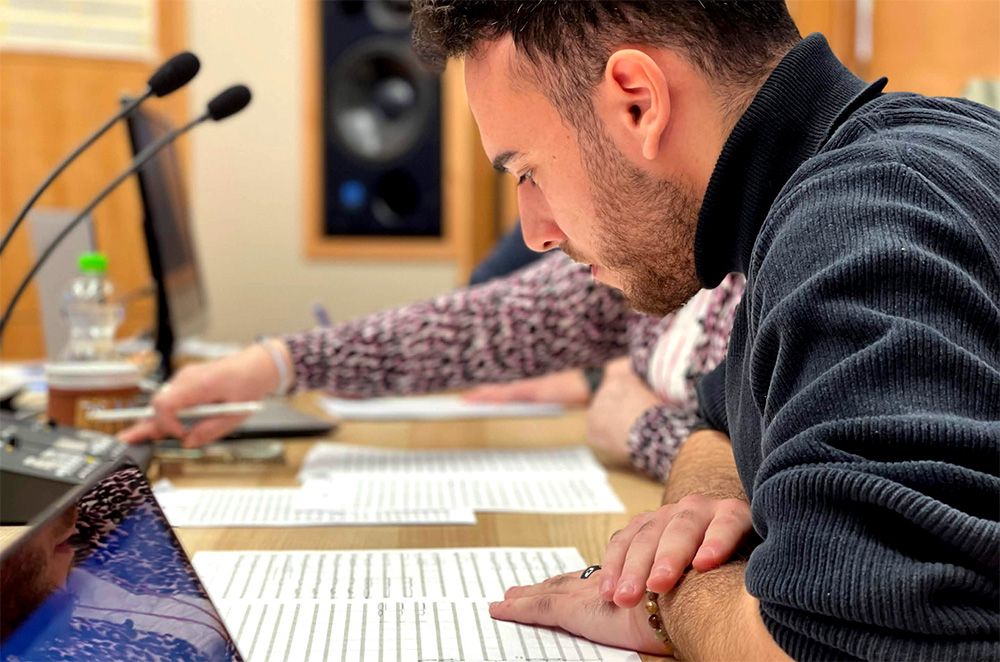 Ein IMAscore-Komponist prüft ausgedruckte Notenblätter an einem Mischpult im Studio, im Hintergrund ein Keyboard und ein Studiomonitor.