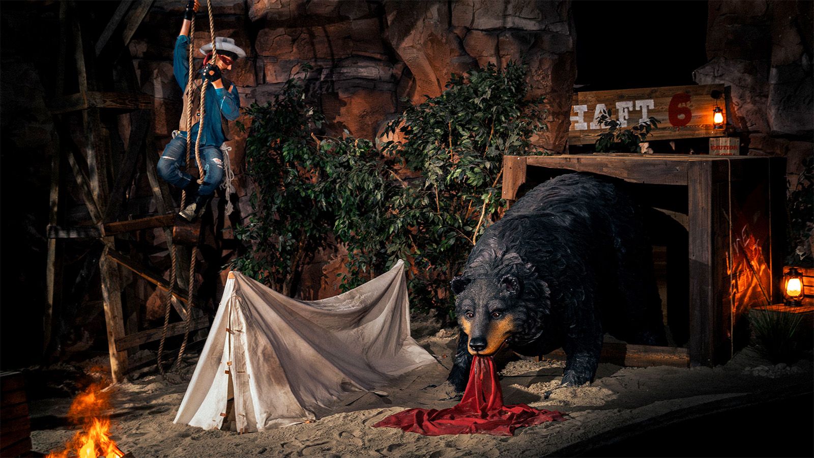 Animatronic bear looming over a torn tent in a themed mine-shaft scene, with a cowboy figure climbing a rope ladder overhead.